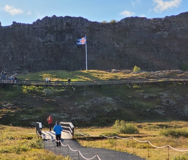 Lögberg séð að austan. Fólk á göngustíg og brú yfir Öxarárhólma og fólk á Lögbergspalli. Blaktandi íslenskur fáni á fánastöng.